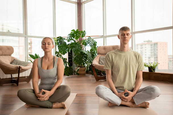 Joven pareja serena en ropa deportiva cruzando sus piernas mientras están sentados en alfombras y practicando ejercicios de meditación juntos en casa - Foto, Imagen