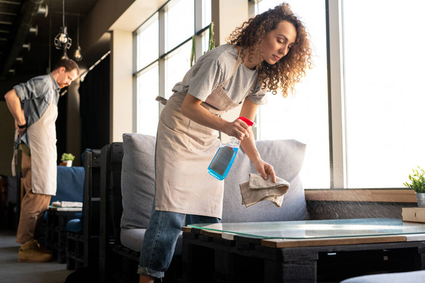 Giovane cameriera dai capelli ricci in grembiule spruzzare detergente mentre bussing tavoli in caffè prima di aprire - Foto, immagini