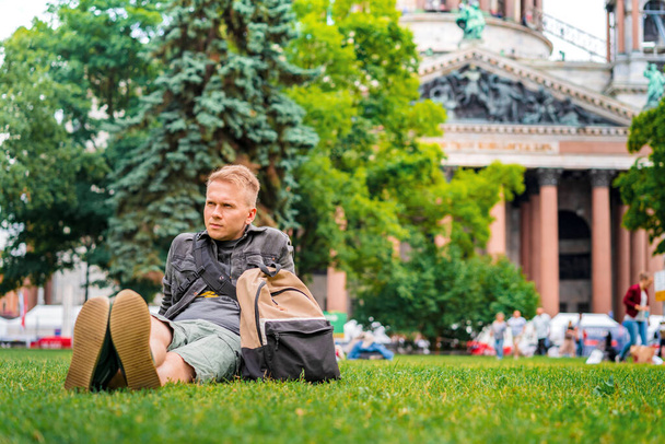 Um jovem turista com uma mochila visita os principais pontos turísticos de São Petersburgo, deitado na grama em frente à Catedral de São Isaac
 - Foto, Imagem