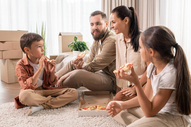 Ystävällinen nuori perhe istuu matolla uudessa talossa täynnä muuttolaatikoita ja syö pizzaa illallisella. - Valokuva, kuva