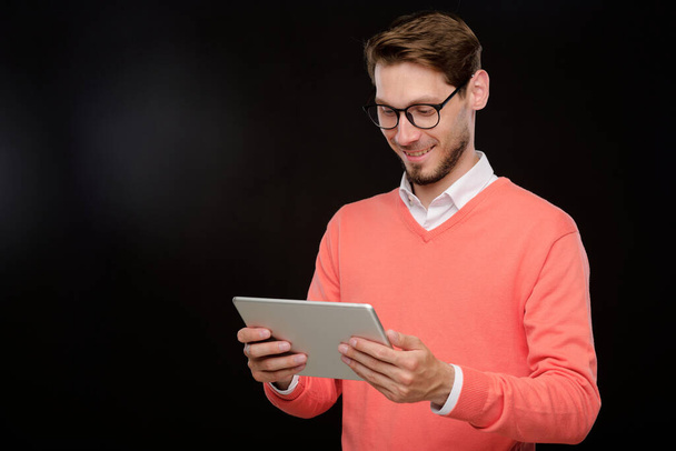 Lächelnder schöner junger bärtiger Mann im hellen Pullover surft vor isoliertem Hintergrund auf einem digitalen Tablet in den sozialen Medien - Foto, Bild