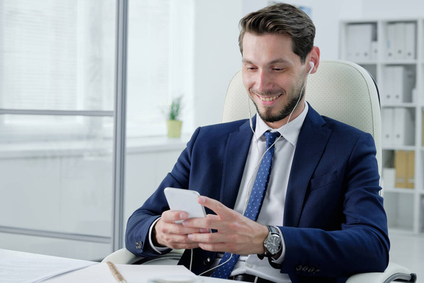 Positive young bearded businessman in earphones sitting on office chair and checking phone while listening to music - Photo, Image
