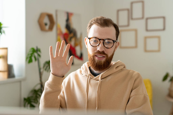 Smiling young freelance designer with beard waving hand while greeting customer via video chat - Photo, Image