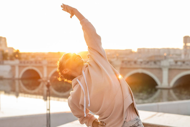 Glückliche junge, fitte Frau in Kapuzenpulli, die ihren Arm ausstreckt und ihn ansieht, während sie im städtischen Umfeld auf dem Hintergrund der Brücke Sport treibt - Foto, Bild