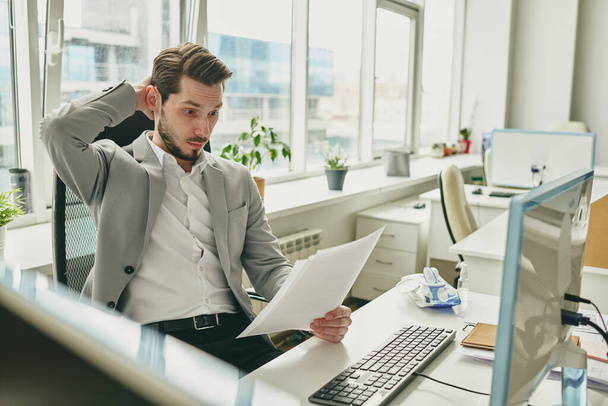 Mladý zmatený podnikatel drží ruku na hlavě, zatímco se dívá na finanční papíry během pracovního dne před počítačem v kanceláři - Fotografie, Obrázek