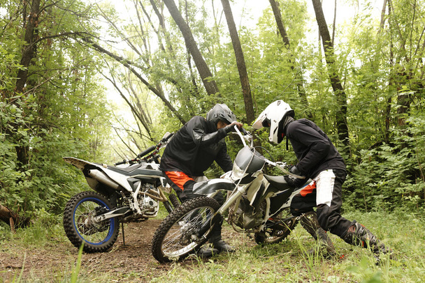 Strong men in helmets picking up motorcycle together in forest while practicing off-road skills - Photo, Image