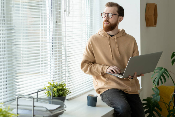 Töprengő, fiatal szakállas férfi kapucnis pulcsiban ül az ablakpárkányon és laptopot tart, miközben a tervezésen gondolkodik. - Fotó, kép