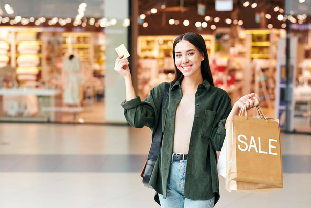 Fiatal vidám vásárló papírzacskókkal mutatja a hitelkártyáját, és nézi, miközben áll a háttérben a bevásárlóközpont osztályok - Fotó, kép