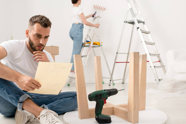 Ciddi genç sakallı adam bacak bacak üstüne atarak oturuyor ve yeni dairede mobilya montajı yaparken ders okuyor. - Fotoğraf, Görsel