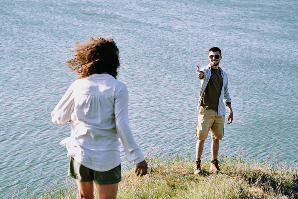 Lächelnder junger männlicher Wanderer streckt seiner Frau den Arm entgegen und lädt sie ein, näher zu kommen und an sonnigen Tagen einen herrlichen Blick auf den See zu genießen - Foto, Bild