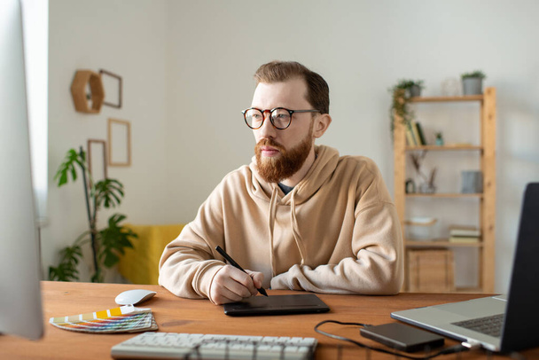 Busy hipster cartoonist with beard sitting at wooden desk and using drawing tablet while making pictures - Fotografie, Obrázek