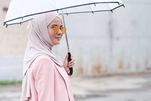 Introspectieve jonge moslimvrouw in roze hijab staande tegen moskee met helder ontwerp en kijkend naar afstand - Foto, afbeelding