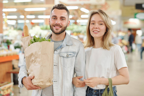 Portrait of smiling beautiful young couple standing with paper bag at farmers market - Fotografie, Obrázek