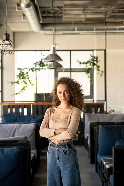 Portrait of content curly-haired entrepreneur standing with crossed arms in own cozy cafe - Fotoğraf, Görsel