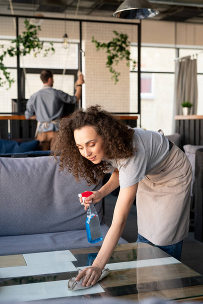 Content curly-haired girl in apron wiping coffee table with cloth while disinfecting surfaces in cafe - Foto, afbeelding