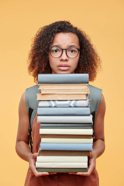 Portrait of scared black student girl in eyeglasses confused with workload holding heap of textbooks against yellow background - Valokuva, kuva