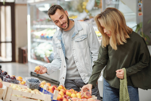 Conteúdo jovem casal em trajes casuais em pé no balcão de alimentos e à procura de frutas maduras no mercado de agricultores - Foto, Imagem
