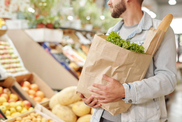 Gros plan de l'homme barbu en veste de denim transportant un sac en papier plein de produits frais sur le marché alimentaire - Photo, image