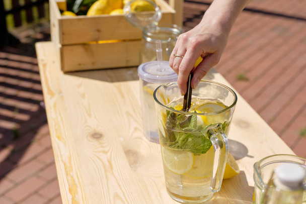 Рука молодої жінки, що змішує інгредієнти домашнього лимонаду в скляному глечику, стоячи на дерев'яній підставці на відкритому повітрі
 - Фото, зображення