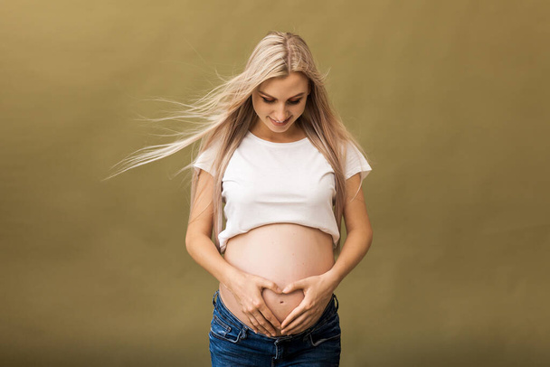 Pregnant happy Woman touching her belly. Pregnant mother portrait, caressing her belly and smiling. Healthy Pregnancy concept, blond expectant female on beige background - Photo, Image