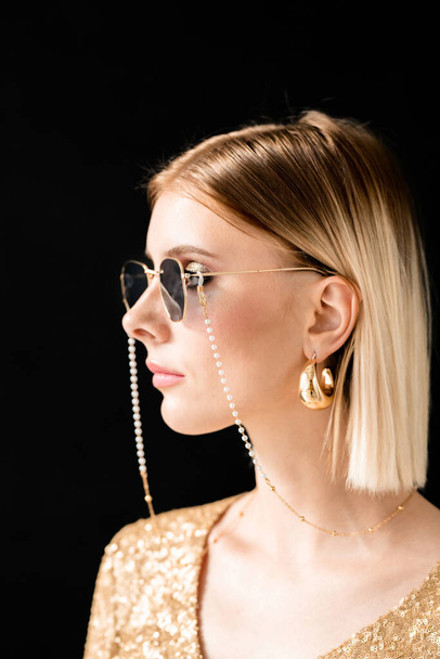 Fiatal szőke, elbűvölő nő stílusos napszemüvegben, csillogó ruhában és arany fülbevalóval a kamera előtt - Fotó, kép