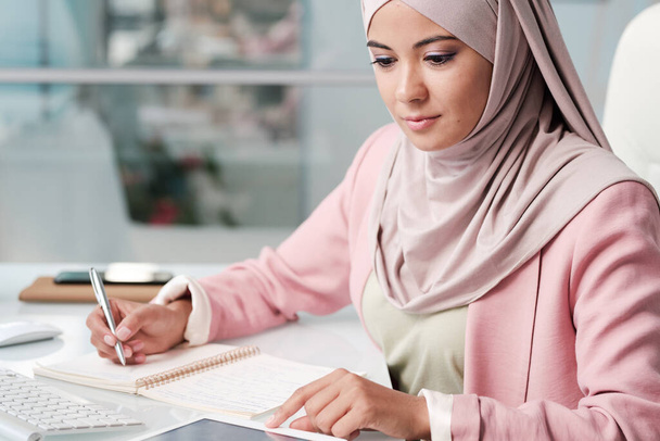 красивая молодая мусульманская бизнес-леди работает на ноутбуке и смотрит в сторону - Фото, изображение