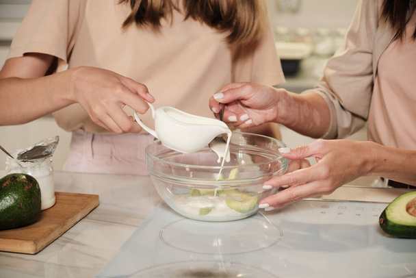 Fiatal nő és lánya kezei, akik friss tejet öntenek az üvegtálba avokádó szeletekkel, miközben házi készítésű jégkrémet készítenek. - Fotó, kép