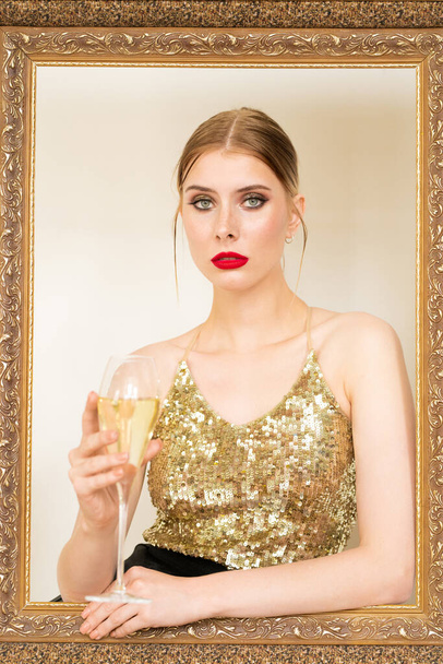 Młoda elegancka blondynka z fletem szampana siedzi za dużą dekoracyjną ramą i robi tosty urodzinowe - Zdjęcie, obraz
