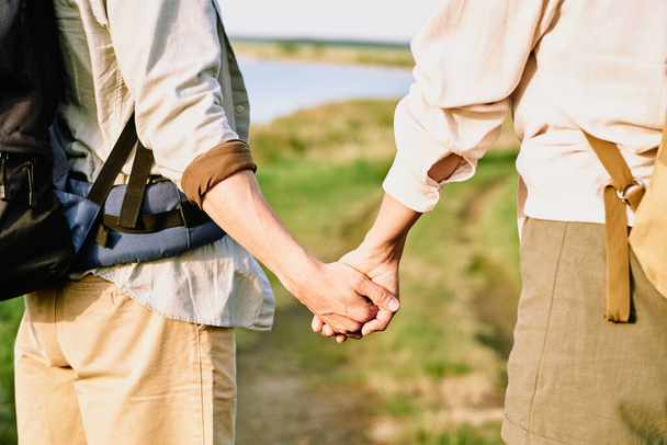 Ruce mladých láskyplný pár s batohy stojící na venkovské silnici, zatímco mají výlet nebo pěší turistiku v přírodním prostředí u jezera - Fotografie, Obrázek