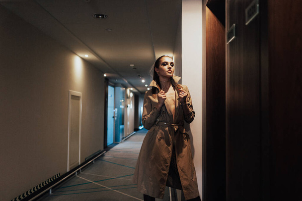 a young beautiful girl, blonde, business lady with bright eyes and a beautiful smile in a long light raincoat stands in the long corridor of the hotel in the light from the duty lamps - Φωτογραφία, εικόνα