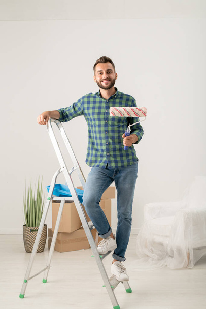 Retrato de joven barbudo alegre de pie con rodillo de pintura en escalera en nuevo apartamento con cosas en movimiento - Foto, imagen