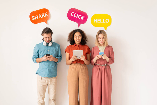 Zwei interkulturelle Mädchen und ein junger Mann in Freizeitkleidung scrollen in mobilen Geräten, während sie in sozialen Netzwerken miteinander kommunizieren - Foto, Bild