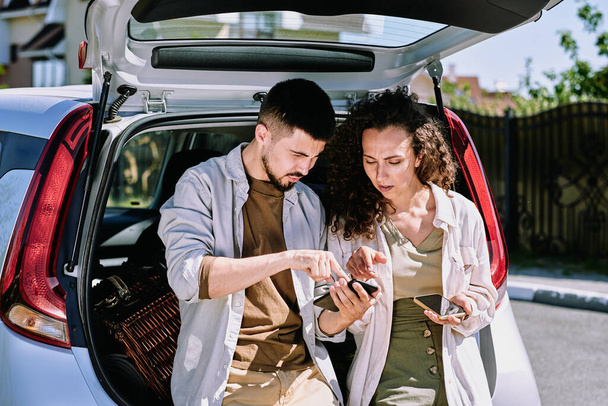 Young man pointing at map in smartphone while explaining his wife where they are going to move and showing her place to stop for rest - Photo, Image