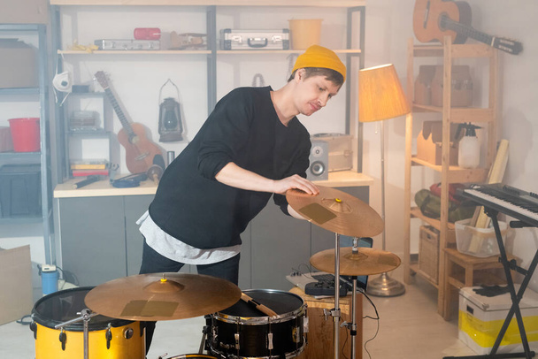 Сучасний молодий музикант у повсякденному одязі нахиляється над барабанним набором, перевіряючи один з цимбалів перед індивідуальною репетицією в гаражі
 - Фото, зображення