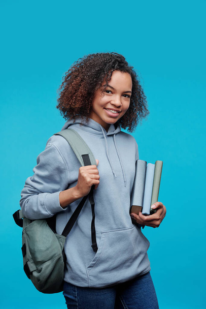 Portrét usmívající se kudrnaté afro-americké studentky stojící s knihami a brašnou na modrém pozadí - Fotografie, Obrázek