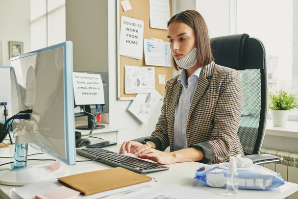 Jovem empresária séria com máscara têxtil protetora no queixo pressionando botões do teclado do computador enquanto trabalhava no escritório - Foto, Imagem