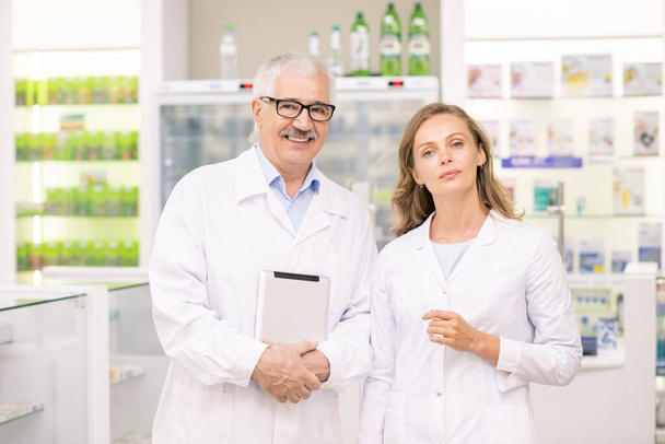Όμορφη ξανθιά γυναίκα φαρμακοποιός και ο ανώτερος συνάδελφός της σε whitecoats στέκεται μπροστά από την κάμερα κατά μεγάλες οθόνες με ιατρική - Φωτογραφία, εικόνα