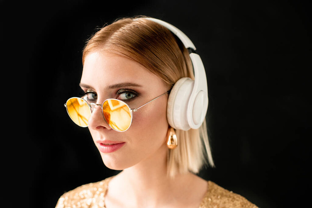 Jovem mulher loira glamourosa em fones de ouvido brancos e óculos de sol elegantes em pé na frente da câmera contra fundo preto - Foto, Imagem