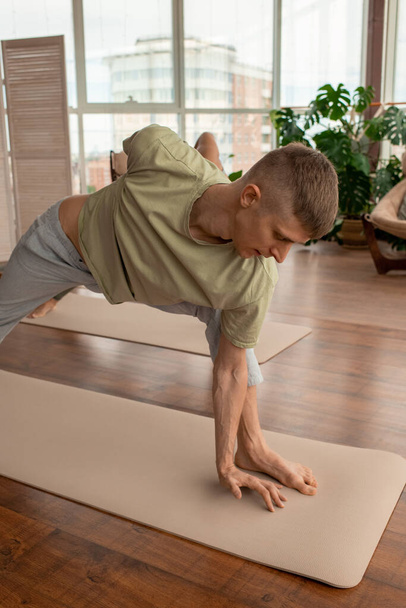Joven hombre activo en ropa deportiva de pie en la estera con un brazo detrás de la espalda mientras practica ejercicios de yoga en el entorno doméstico - Foto, imagen