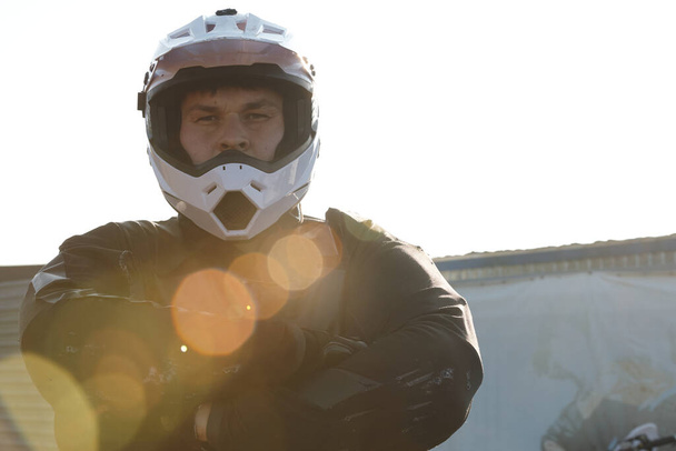 Koruyucu giysili, kasklı, kolları çaprazlanmış, güneş ışığı efektli ciddi bir motorsikletçinin portresi. - Fotoğraf, Görsel