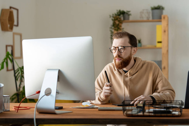 Skoncentrowany młody brodaty projektant w okularach i kapturze siedzi przy biurku i analizuje szkice na komputerze - Zdjęcie, obraz
