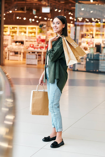 Cliente femenino bastante joven con bolsas de papel de pie delante de la cámara dentro de un gran centro comercial moderno durante la venta estacional - Foto, imagen