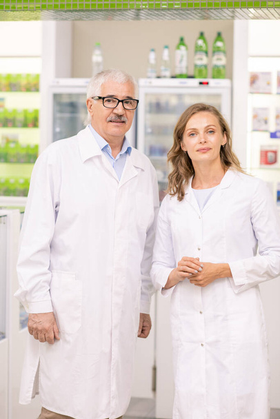 Two successful professionals in whitecoats standing between large display with medicines and counter inside contemporary drugstore - Photo, Image