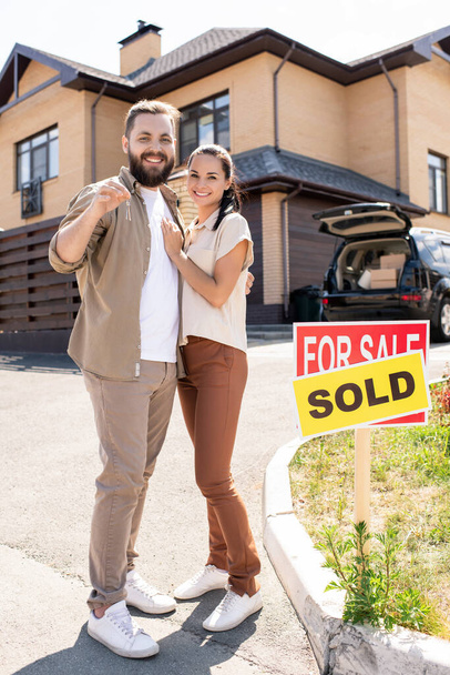 Portrait of happy beautiful young couple in casual outfits standing against new house and embracing each other while showing key from new house - Photo, Image