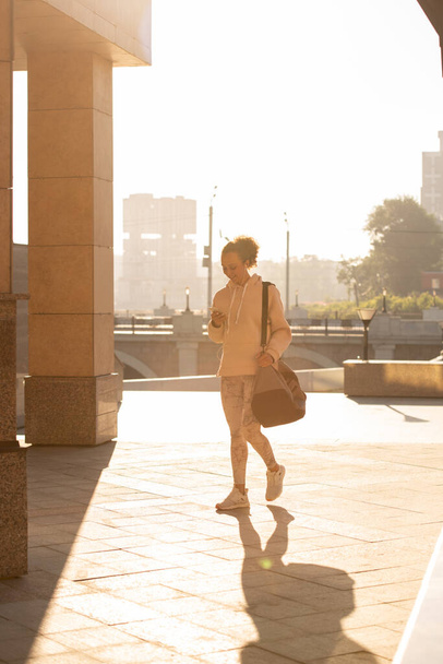 Fiatal, aktív sportos nő táskával a vállán görgetés okostelefonon, miközben hazajön szabadtéri edzés után a napsütéses napon - Fotó, kép