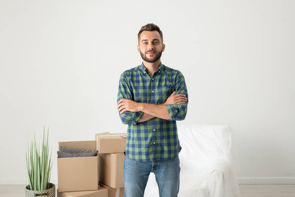 Retrato de joven guapo contenido en camisa a cuadros de pie con brazos cruzados en nuevo piso con cajas móviles - Foto, Imagen