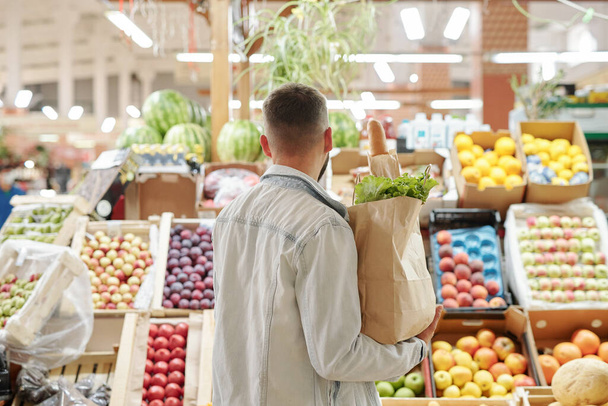 Açık renk kot ceketli genç adam elinde kese kağıdıyla çiftçi pazarından sulu meyveler seçiyor. - Fotoğraf, Görsel