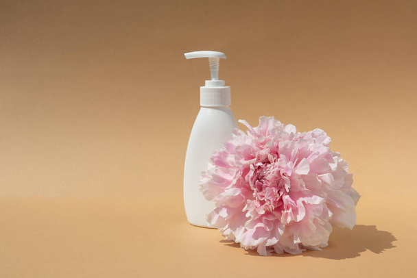 white cosmetic bottle and pink peony bud on brown background. Skin hydration and health - Photo, image