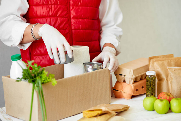 Vrijwilliger in de beschermende medische masker en handschoenen zetten voedsel in donatie doos. Leveringsman werknemer in rode vest verpakking doos met voedsel. Bedrijfsquarantaine pandemisch coronavirus. - Foto, afbeelding