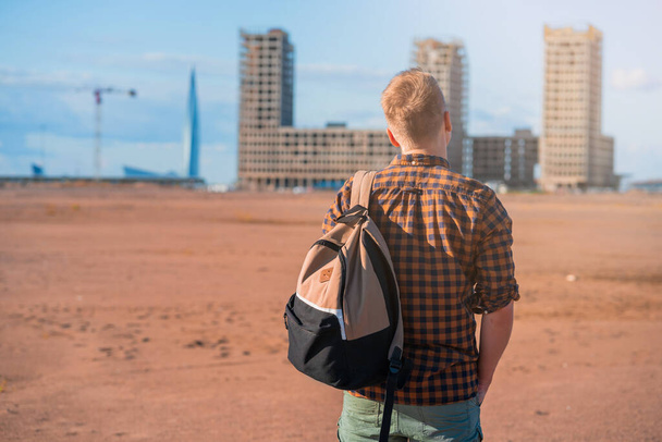  fiatal férfi ellenőrzött ingben, hátizsákkal egy üres telken áll, több emeletes épületek előtt, a településre várva. - Fotó, kép
